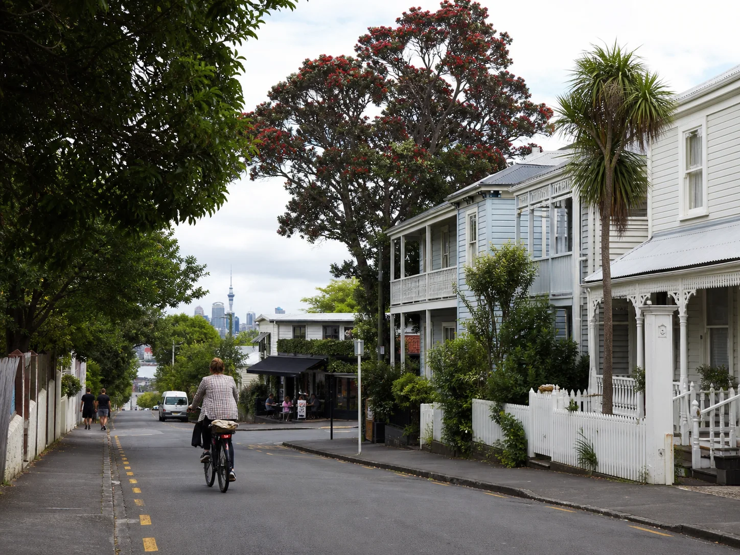 Leafy Grey Lynn street with timber villas, a corner café, a cyclist and the Auckland skyline in the distance