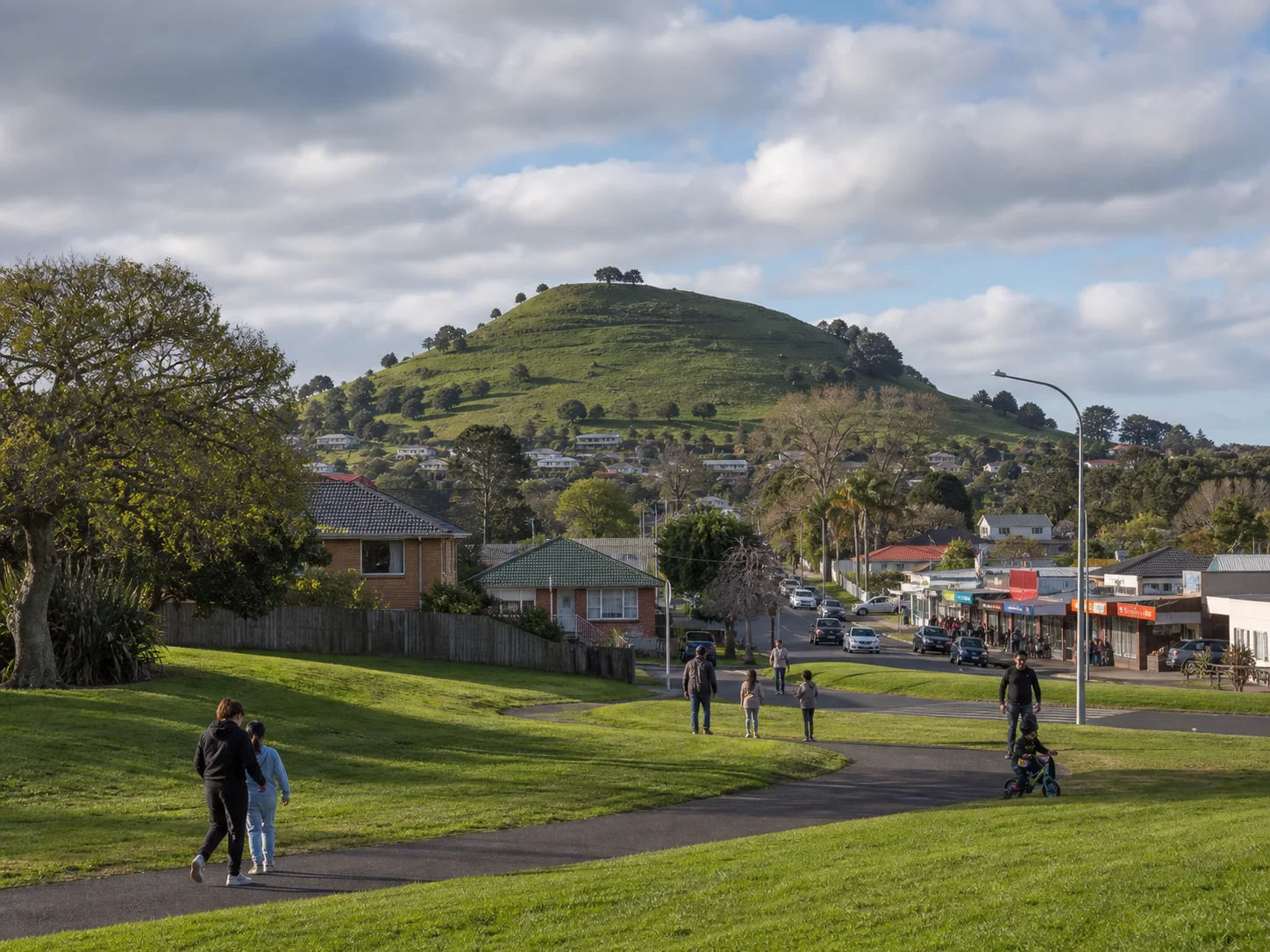 Puketāpapa rising above a Mount Roskill reserve, local shops and families walking through the neighbourhood